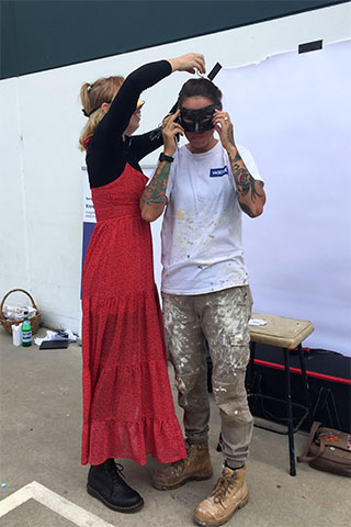 A person in a long red dress and black long-sleeved t-shirt tying a black masquerade mask around the face of another person in a worn white t-shirt, khaki workpants splattered with paints, and work boots. In the background a white backdrop can be seen taped to a wall with a stool placed in front of it.