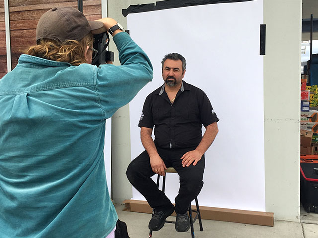 Duncan Wright shooting a portrait of a middle aged person in a short black shirt, black trousers, and black trainers, sitting at a make-shift studio setup.