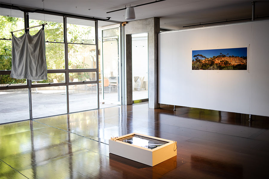 A wide view of a well-lit room, with a textile work hanging from the ceiling on the left and a wall with a landscape photograph on the right. In the middle of the room is a white box on the ground with a reflective surface.