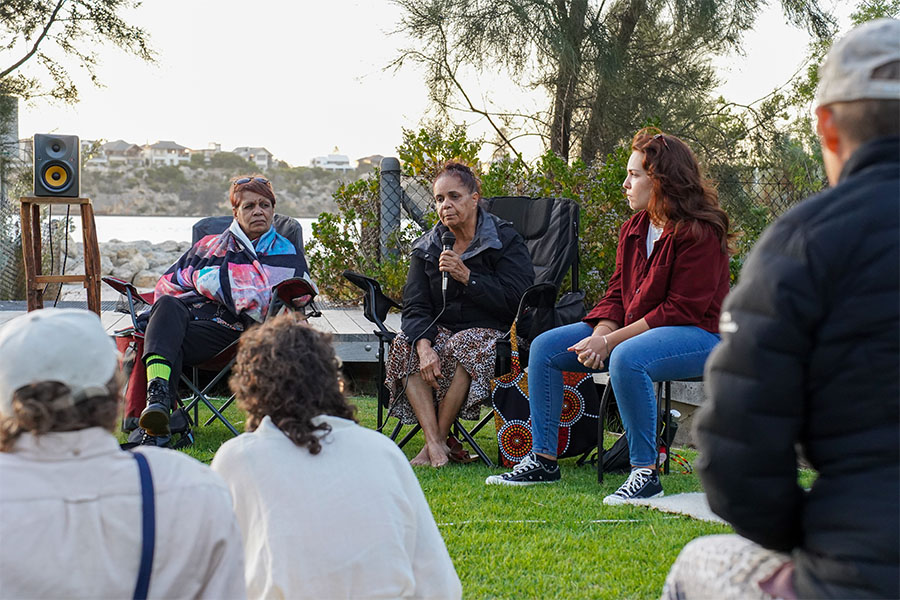 A group of three people are seated on chairs in a grassy area. The person in the middle is holding a microphone, with the person on the left looking at them and artist Yabini Kickett on the right looking into the distance while listening to them. There is a view of the river behind the group, and they are addressing an audience seated on the ground.