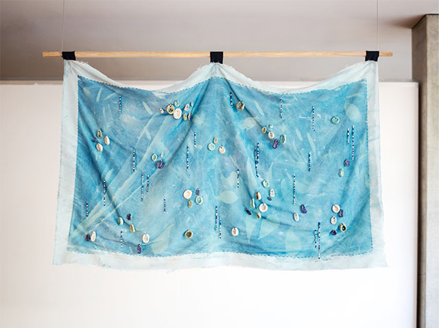 Full view of the textile work hanging at UWA Cullity Gallery. The cloth is dyed light blue–the inner section framed by a faded blue section. The separation between the two sections is emphasized by dark blue darning. Various leaf shapes can be seen in silhouetted form. Small clay and beaded string charms are sewn on to the fabric. The work is attached to a hanging wood dowel with three black loops sown to the top.