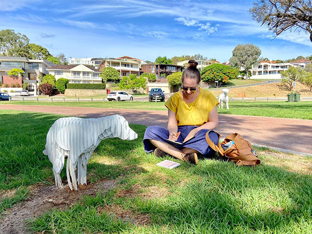Artist Amy Perejuan-Capone is sitting cross-legged on the grass in a park. She is wearing a yellow t-shirt and blue trousers and appears to be writing into a notebook. There is a white sculpture of a dog immediately in front of her, as well as in the distance behind her.
