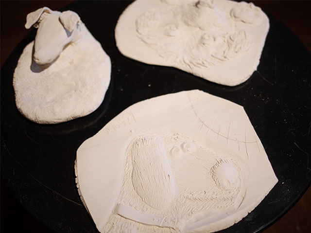Close-ups of three white unglazed ceramic pieces. The one closest to the camera has a raised pattern of a plant on it. To its left is a small sculpture of a dog’s head. The third piece on the right has a less discernible raised pattern.
