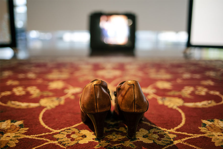 A pair of patinated brown high heel shoes on a red rug patterned with golden flowers and curved lines. The photo is taken fron ground level. A CRT (Cathode Ray Tube) television can be seen in the distance, out of focus.