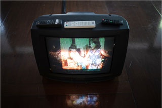 A close-up shot looking down at a black CRT (Cathode Ray Tube) television placed on the floor. Atop the television are three remotes. The television shows a frame of a home video of two young girls.