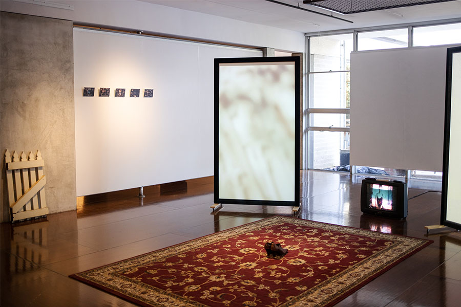 A view of the installation at UWA Cullity Gallery. In the center of the room is a red rug patterned with golden flowers and curved lines. Placed on top of it is a pair of brown leather high heels facing a small black CRT (Cathode Ray Tube) television playing a home video. Flanking the television are two tall framed white screens with videos projected on them. On left wall surrounding the exibit are five small photos —&thisp;still frames from a video. Towards the left edge of the photo, a white picket gate can be seen leaned against the wall.