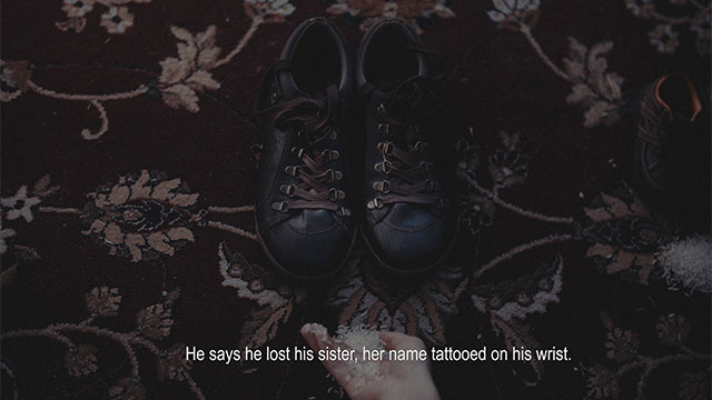 A still frame of a video work by the artist tiled 'our home'. The frame is a top-down view of a handful of rice being placed in front of a pair of dark brown leather boots. The boots are placed atop a red rug patterned with golden flowers and curved lines. A caption reads 'He says he lost his sister, her name tattoed on his wrist.'