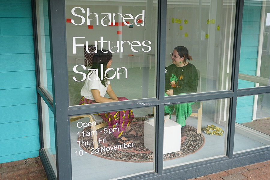 Ana in conversation with another person in a glass room. Text applied on to the glass reads 'Shared Futures Salon'.