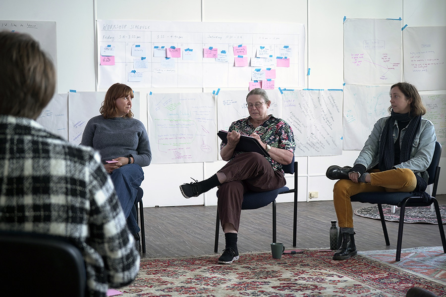 Four people seen seated, in conversation. The person in the center is taking notes. In the background are various drawings, writing, and a schedule stuck to the wall.