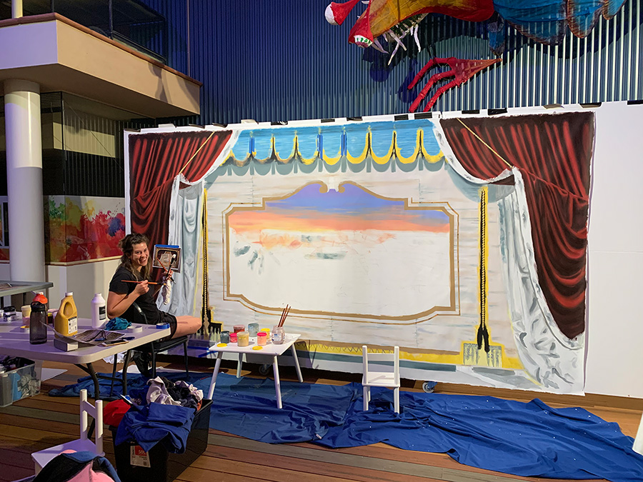 Artist Georgie Mattingley painting a large scale backdrop with a reference image on hand. They are half turned towards the camera, smiling.