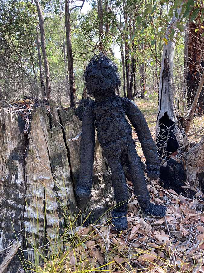 A sculpture resembling both a humanoid form and charred wood leaning against a burnt tree trunk in the bush
