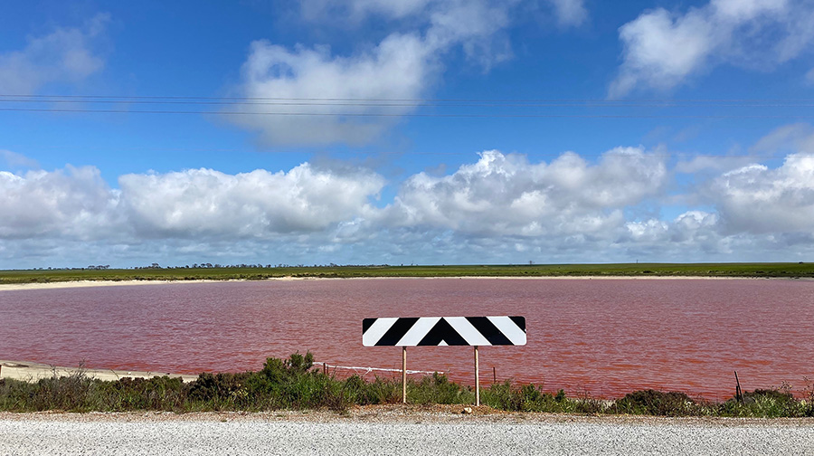 Pink Lake as seen during day time. A black and white, striped, 'No Through Road' sign is in the foreground followed by the lake, and the landscape beyond. The sky is clear closer to the foreground and cloudy towards the horizon