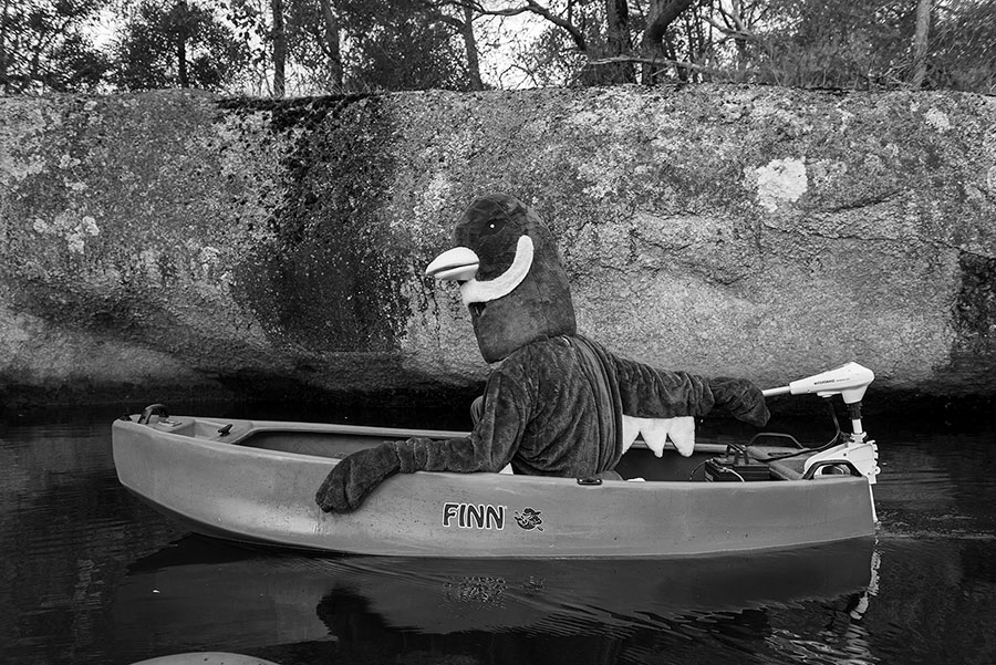 Black and white photo of a person in a bird costume operating a small motor boat