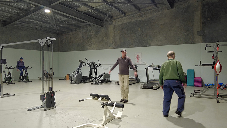 Three elderly men in different states of exercise at a gym. Various exercise equipment are arranged against the walls.