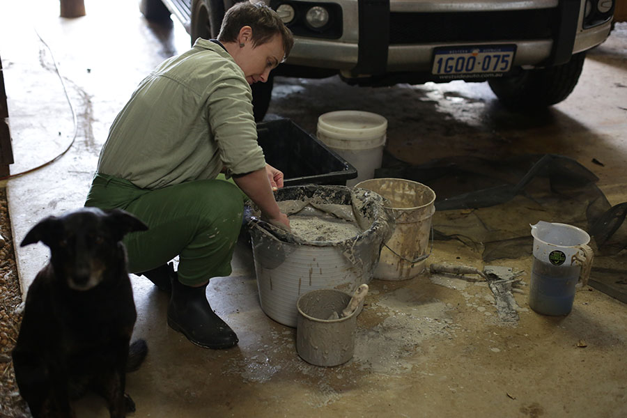 Elizabeth Pedler mixing clay in buckets in the garage. A black doggo can be seen out of focus on the bottom left.